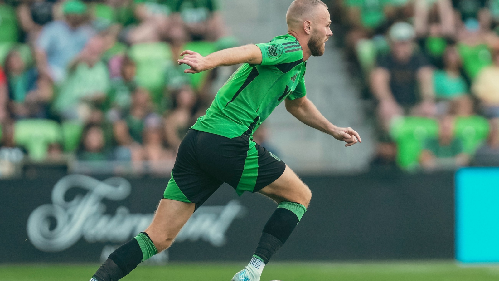 Soccer player in a green and black kit sprinting on the field during a match, blurred crowd in the stands behind.