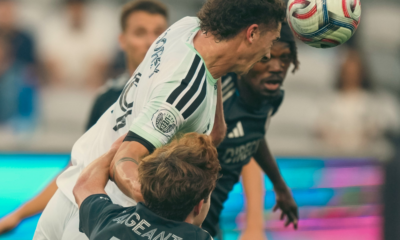 Two soccer players contest a header, one in white jersey and the other in black, as the ball descends toward them.
