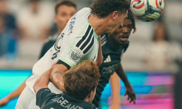 Two soccer players contest a header, one in white jersey and the other in black, as the ball descends toward them.