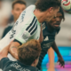 Two soccer players contest a header, one in white jersey and the other in black, as the ball descends toward them.