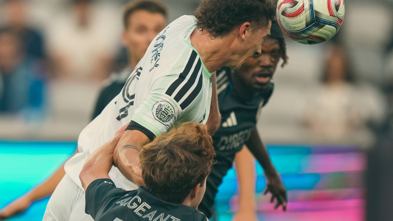 Two soccer players contest a header, one in white jersey and the other in black, as the ball descends toward them.