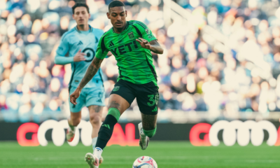 Soccer player in a bright green jersey dribbles the ball on a grassy field with a blurred crowd behind him in the stands.