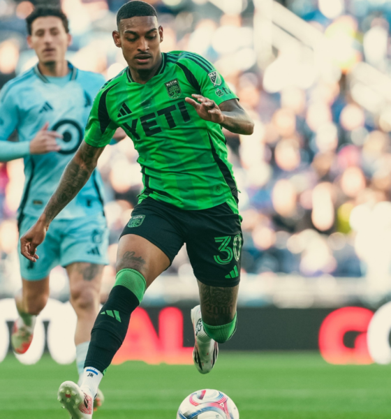 Soccer player in a bright green jersey dribbles the ball on a grassy field with a blurred crowd behind him in the stands.