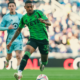 Soccer player in a bright green jersey dribbles the ball on a grassy field with a blurred crowd behind him in the stands.