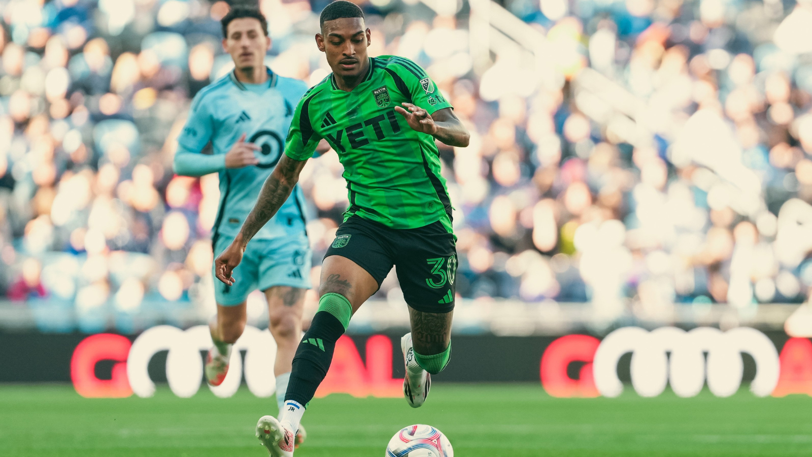 Soccer player in a bright green jersey dribbles the ball on a grassy field with a blurred crowd behind him in the stands.