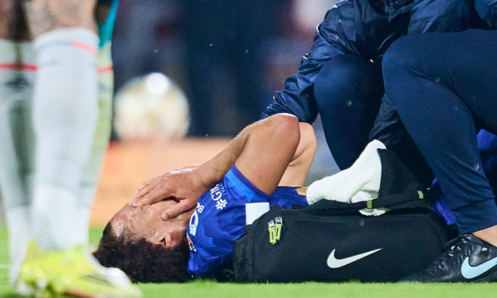 Soccer player in blue on the grass receives medical attention from a trainer beside a black Nike bag.