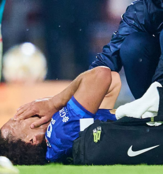 Soccer player in blue on the grass receives medical attention from a trainer beside a black Nike bag.
