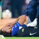 Soccer player in blue on the grass receives medical attention from a trainer beside a black Nike bag.
