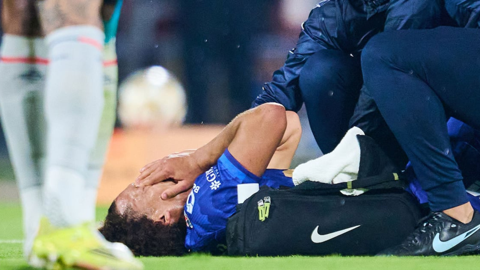 Soccer player in blue on the grass receives medical attention from a trainer beside a black Nike bag.