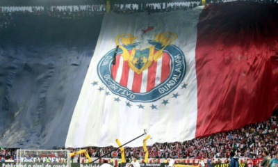 Massive banner flag unfurls across the stadium stands, featuring red-and-white stripes and a circular crest for a soccer club, with fans cheering below.