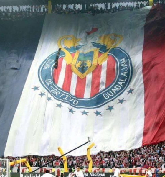 Massive banner flag unfurls across the stadium stands, featuring red-and-white stripes and a circular crest for a soccer club, with fans cheering below.