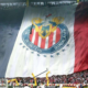 Massive banner flag unfurls across the stadium stands, featuring red-and-white stripes and a circular crest for a soccer club, with fans cheering below.