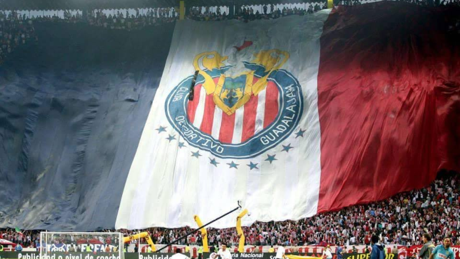 Massive banner flag unfurls across the stadium stands, featuring red-and-white stripes and a circular crest for a soccer club, with fans cheering below.