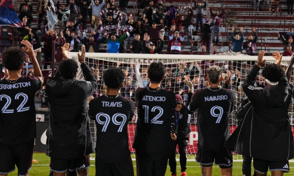 Soccer players with backs to the camera applaud a packed stadium, wearing black kits with names and numbers (Herrington 22, Travis 99, Atencio 12, Navarro 9).