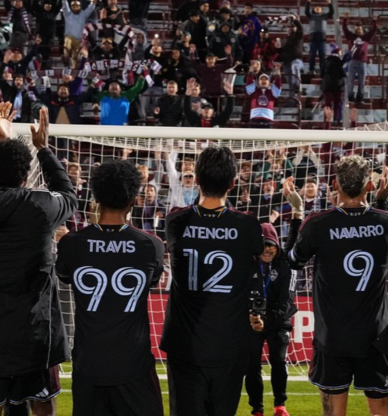 Soccer players with backs to the camera applaud a packed stadium, wearing black kits with names and numbers (Herrington 22, Travis 99, Atencio 12, Navarro 9).