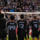Soccer players with backs to the camera applaud a packed stadium, wearing black kits with names and numbers (Herrington 22, Travis 99, Atencio 12, Navarro 9).