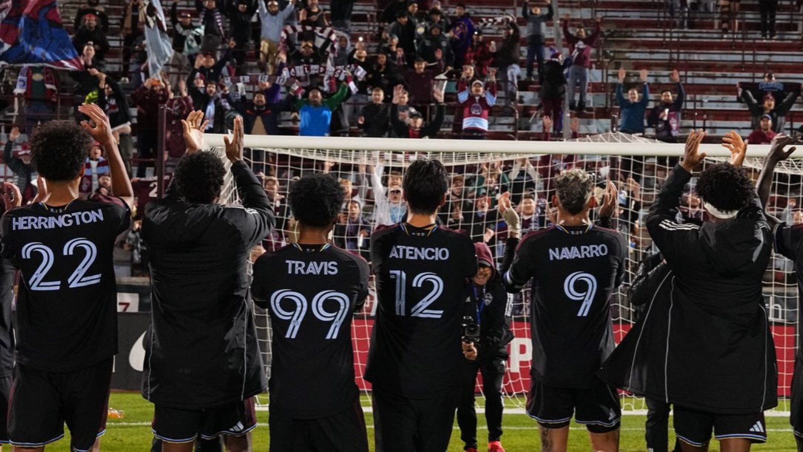 Soccer players with backs to the camera applaud a packed stadium, wearing black kits with names and numbers (Herrington 22, Travis 99, Atencio 12, Navarro 9).