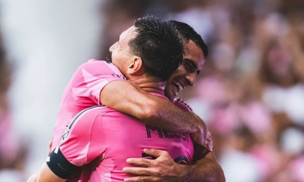 Two teammates in pink jerseys hug in celebration on the soccer field.