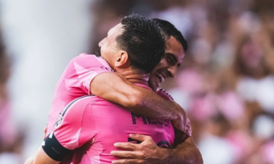 Two teammates in pink jerseys hug in celebration on the soccer field.