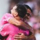 Two teammates in pink jerseys hug in celebration on the soccer field.