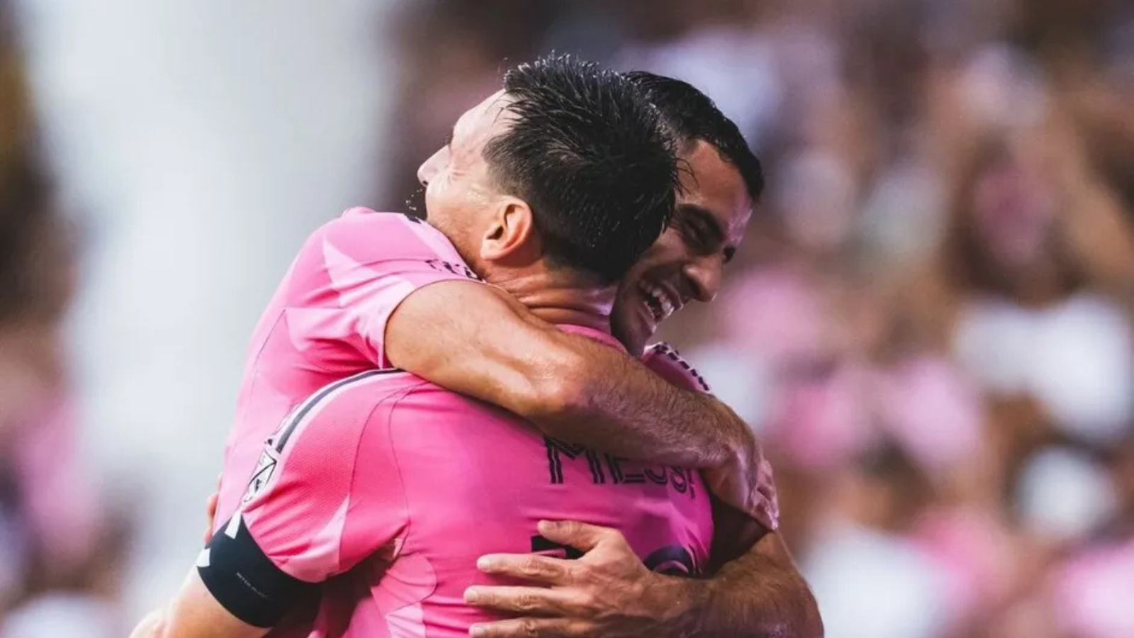 Two teammates in pink jerseys hug in celebration on the soccer field.