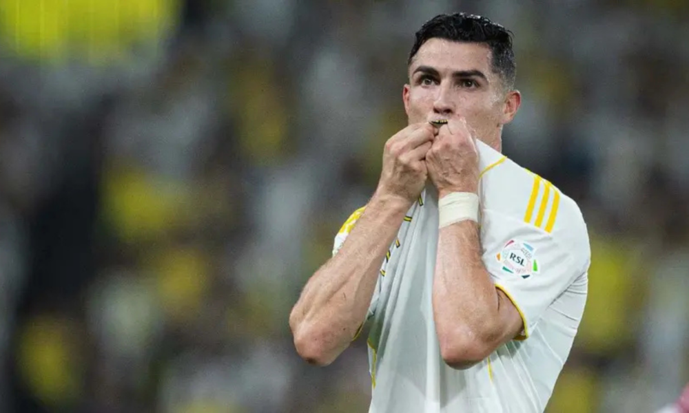 Male soccer player in a white jersey kisses a medal or lanyard after a match, stadium crowd blurred in the background, rain.