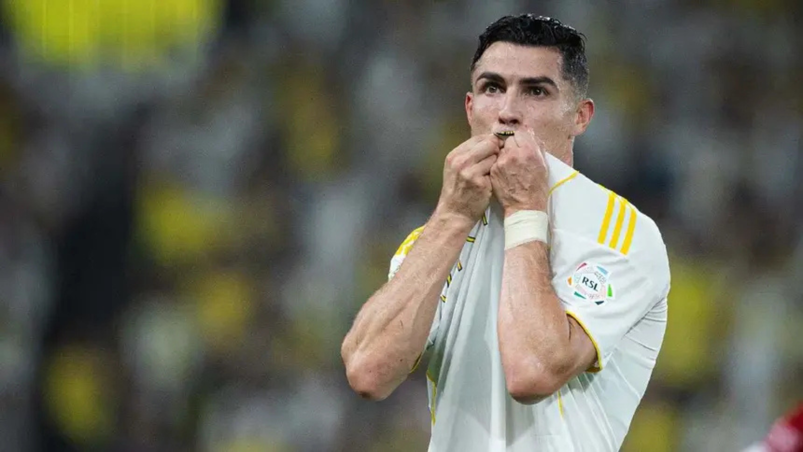 Male soccer player in a white jersey kisses a medal or lanyard after a match, stadium crowd blurred in the background, rain.