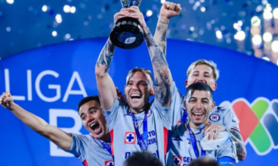 Team of four athletes in light-blue jerseys celebrate on stage, one raising a trophy overhead amid confetti and blue backdrop.
