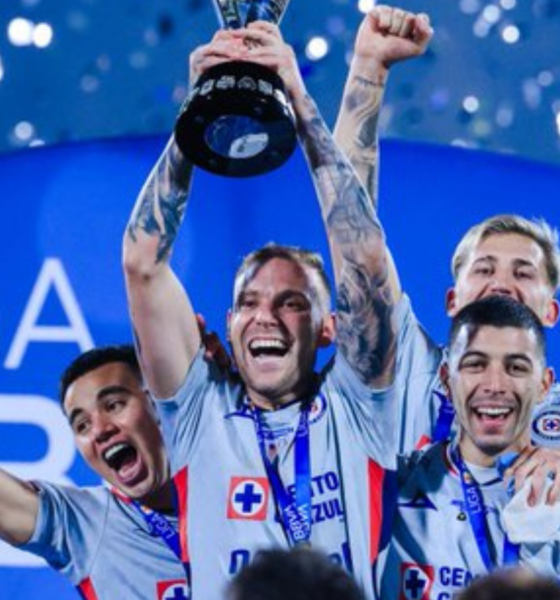 Team of four athletes in light-blue jerseys celebrate on stage, one raising a trophy overhead amid confetti and blue backdrop.