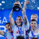 Team of four athletes in light-blue jerseys celebrate on stage, one raising a trophy overhead amid confetti and blue backdrop.
