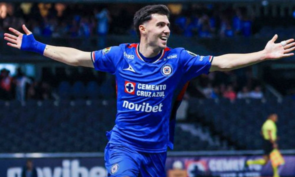 Cruz Azul footballer in a blue kit celebrates with arms outstretched on the field.