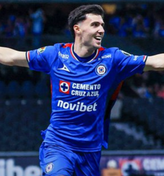 Cruz Azul footballer in a blue kit celebrates with arms outstretched on the field.