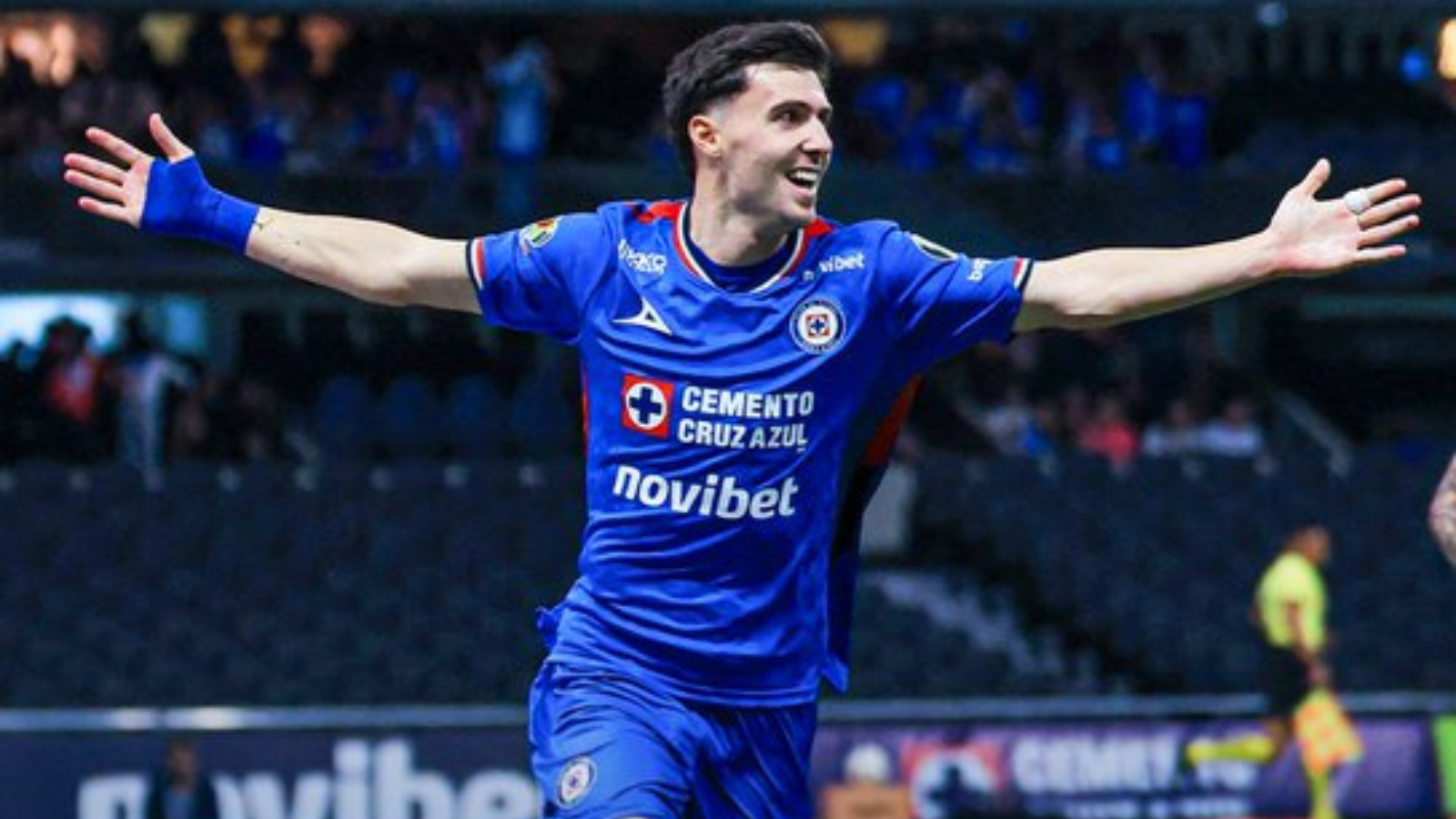 Cruz Azul footballer in a blue kit celebrates with arms outstretched on the field.