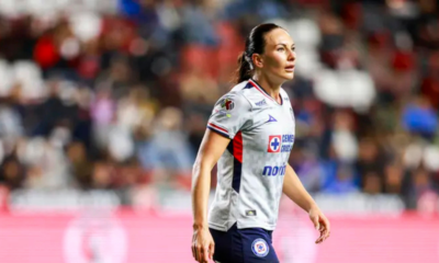 Female soccer player in a white jersey walking on the pitch during a match, blurred stadium crowd in the background.