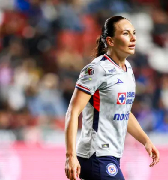 Female soccer player in a white jersey walking on the pitch during a match, blurred stadium crowd in the background.