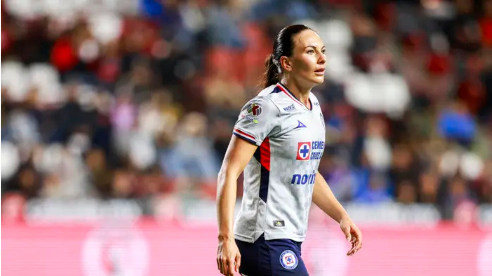 Female soccer player in a white jersey walking on the pitch during a match, blurred stadium crowd in the background.