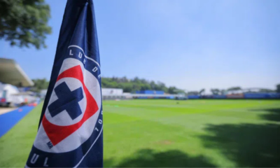 Close-up of a dark blue flag with a red-and-white circular logo in the foreground, a green sports field and blue sky in the background.