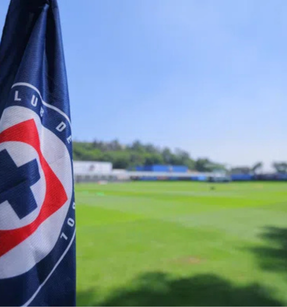Close-up of a dark blue flag with a red-and-white circular logo in the foreground, a green sports field and blue sky in the background.