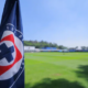 Close-up of a dark blue flag with a red-and-white circular logo in the foreground, a green sports field and blue sky in the background.