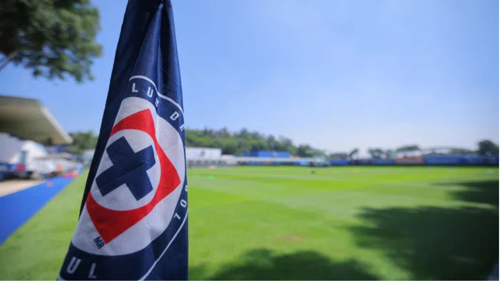 Close-up of a dark blue flag with a red-and-white circular logo in the foreground, a green sports field and blue sky in the background.