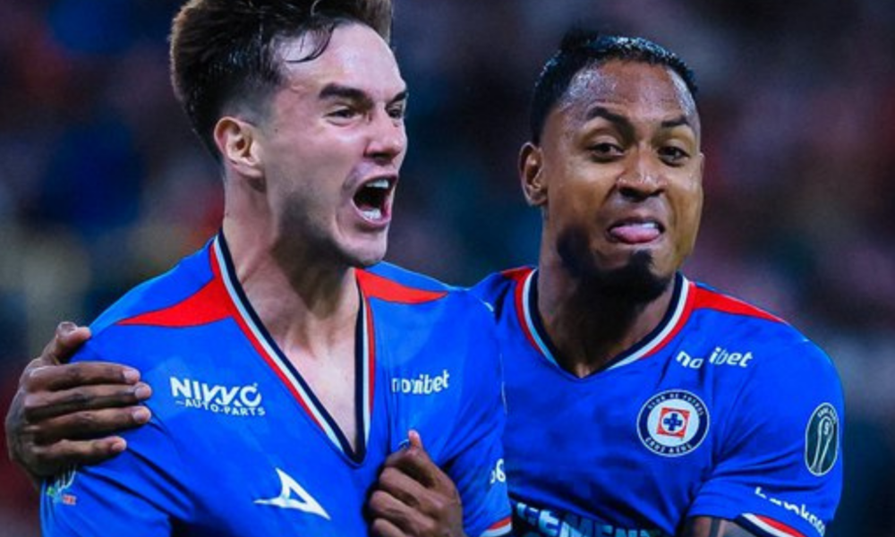 Two soccer players in blue jerseys celebrate together on the field, one with an arm around the other’s shoulder.