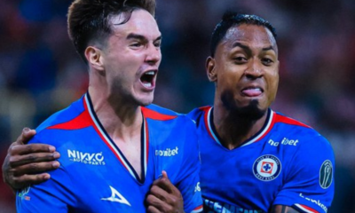 Two soccer players in blue jerseys celebrate together on the field, one with an arm around the other’s shoulder.