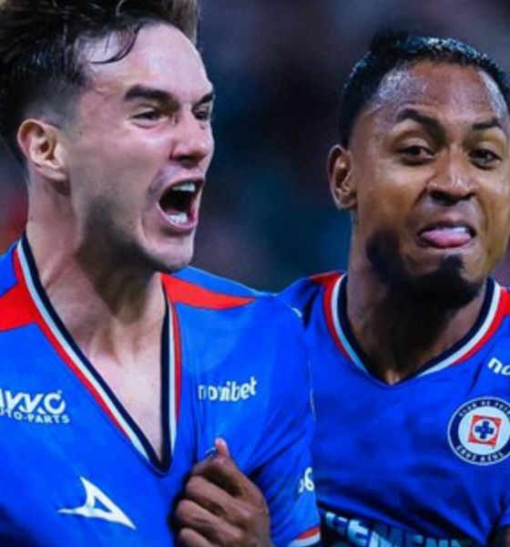 Two soccer players in blue jerseys celebrate together on the field, one with an arm around the other’s shoulder.