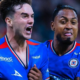 Two soccer players in blue jerseys celebrate together on the field, one with an arm around the other’s shoulder.
