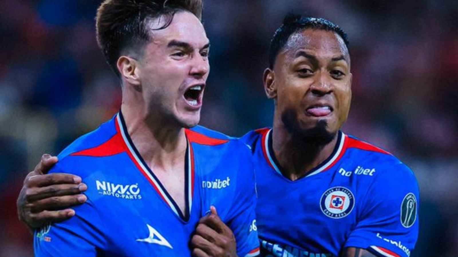 Two soccer players in blue jerseys celebrate together on the field, one with an arm around the other’s shoulder.