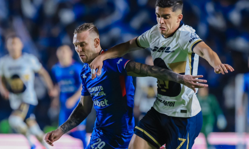 Two soccer players clash for the ball during a night match, blue kit against white with yellow accents, blurred crowd in the background.