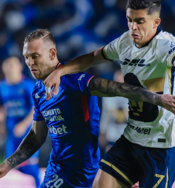 Two soccer players clash for the ball during a night match, blue kit against white with yellow accents, blurred crowd in the background.