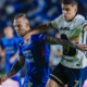 Two soccer players clash for the ball during a night match, blue kit against white with yellow accents, blurred crowd in the background.