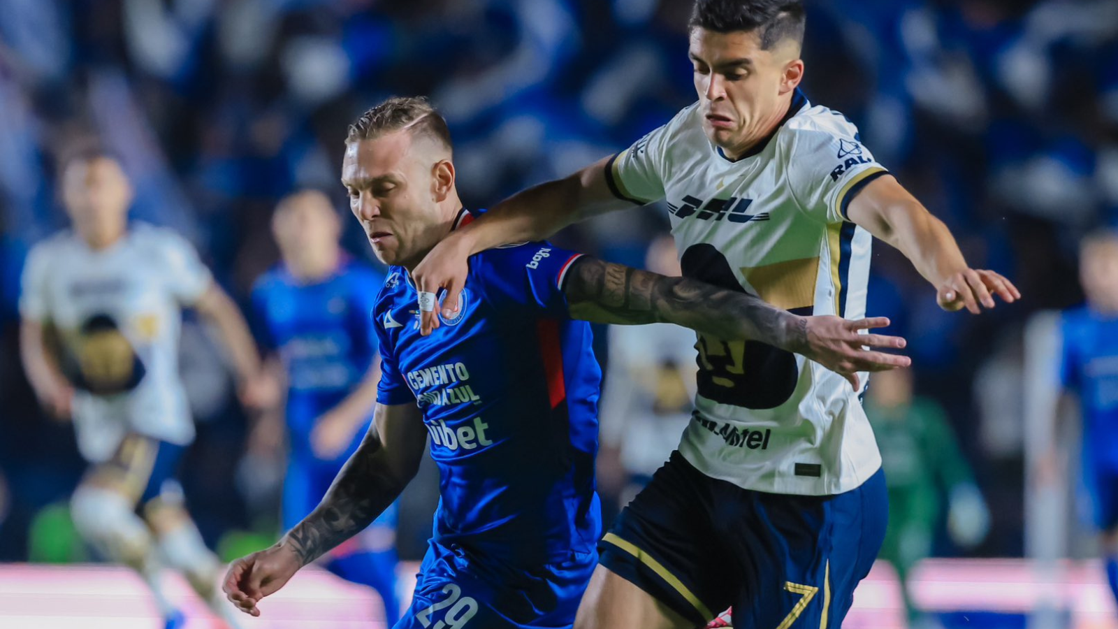 Two soccer players clash for the ball during a night match, blue kit against white with yellow accents, blurred crowd in the background.