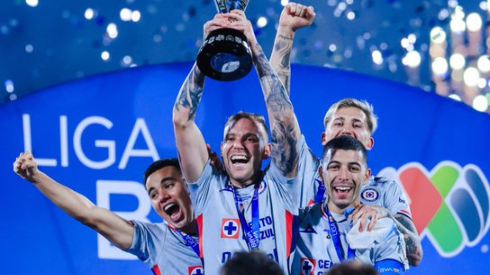 Team of four athletes in light-blue jerseys celebrate on stage, one raising a trophy overhead amid confetti and blue backdrop.